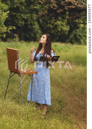 Beautiful young woman is relaxing while painting an art canvas outdoors in the park at rain. 104095677