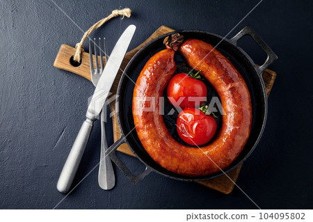 fried meat sausage in a cast iron skillet with tomatoes and cutlery on a wooden cutting board on a dark stone table fried meat sausage in a cast iron skillet with tomatoes and cutlery on a wooden cutting board on a dark stone table 104095802