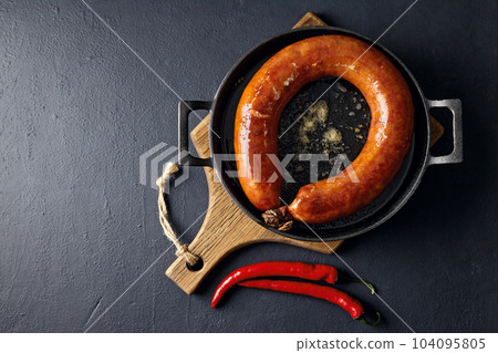 fried meat sausage in a cast iron pan and hot chili pepper on a cutting wooden board on a dark stone table fried meat sausage in a cast iron pan and hot chili pepper on a cutting wooden board on a dark stone table 104095805