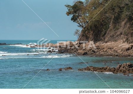 Beautiful view of waves splashing on rocky beach and trees growing on cliff with clear blue sky in background during sunny day at Costa Rica, nature and travel concept Beautiful view of waves splashing on rocky beach and trees growing on cliff with clear blue sky in background during sunny day at Costa Rica, nature and travel concept 104096027