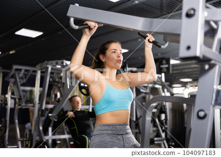 Active young woman training at shoulder press machine in gym 104097183