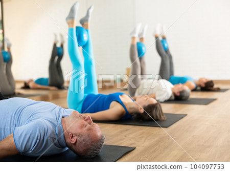 Class of people of different ages holding balls between legs and pushing press during Pilates training in studio Class of people of different ages holding balls between legs and pushing press during Pilates training in studio 104097753