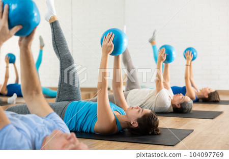 Class of male and women of different ages in active wear pressing balls in hands exercise during Pilates training together in gym Class of male and women of different ages in active wear pressing balls in hands exercise during Pilates training together in gym 104097769