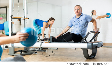 Focused aged man training on reformer equipment in rehabilitation center 104097842