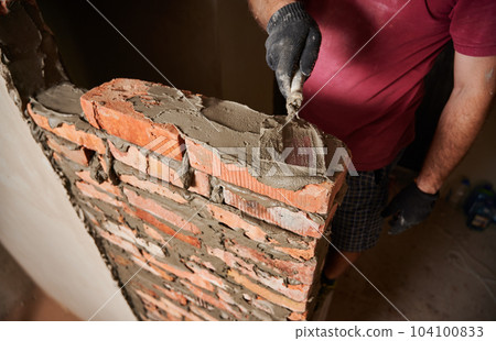Close up of male hands in work gloves laying brickwork in building under construction. Man bricklayer builder applying cement mortar on bricks with trowel tool. Masonry construction concept. Close up of male hands in work gloves laying brickwork in building under construction. Man bricklayer builder applying cement mortar on bricks with trowel tool. Masonry construction concept. 104100833