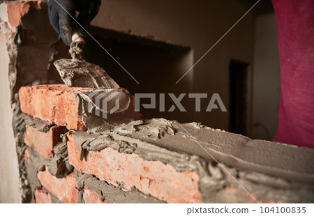 Close up of man hand in work glove laying brickwork in building under construction. Male worker applying cement mortar on bricks with trowel tool. Masonry construction concept. Close up of man hand in work glove laying brickwork in building under construction. Male worker applying cement mortar on bricks with trowel tool. Masonry construction concept. 104100835