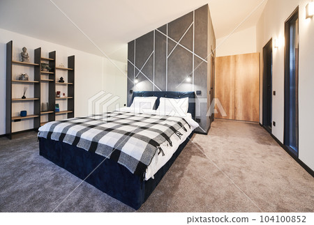 Simply furnished sleeping area with big bed and wooden shelves with buddhist decoration at home. Side view of dark bed covered with plaid bedspread against gray wall in bedroom. Concept of interior. Simply furnished sleeping area with big bed and wooden shelves with buddhist decoration at home. Side view of dark bed covered with plaid bedspread against gray wall in bedroom. Concept of interior. 104100852