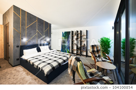 Cozy modern furnished chalet room with reflection in large panoramic window at twilight. Side view of illuminated bedroom indoors. Concept of interior. Cozy modern furnished chalet room with reflection in large panoramic window at twilight. Side view of illuminated bedroom indoors. Concept of interior. 104100854