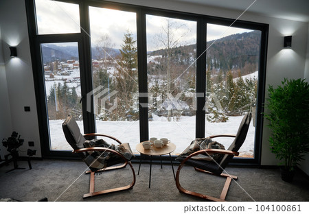 Cozy room with small coffee table and armchairs in front of large panoramic window. Front view of two wooden chairs and table against wide window with snowy mountain landscape. Concept of holidays. Cozy room with small coffee table and armchairs in front of large panoramic window. Front view of two wooden chairs and table against wide window with snowy mountain landscape. Concept of holidays. 104100861