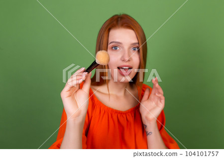 Young red hair woman in casual orange blouse on green background perfect natural smooth skin holding blush brush 104101757