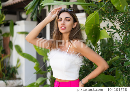 Stylish fit tanned beautiful woman in sunglasses, fashion pink pants and white top posing outdoor at luxury tropical villa 104101758