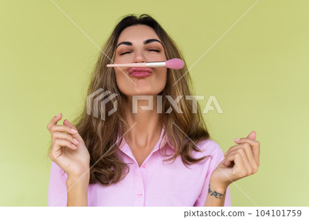 Young woman in casual wear isolated on green background playful positive with blush brush, natural daily makeup Young woman in casual wear isolated on green background playful positive with blush brush, natural daily makeup 104101759