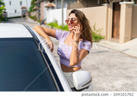Successful smiling fit tanned attractive woman in casual wear is using her smart phone while standing near luxury modern car outdoors. Successful smiling fit tanned attractive woman in casual wear is using her smart phone while standing near luxury modern car outdoors. 104101762