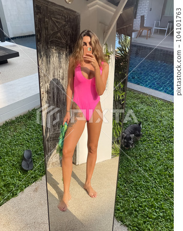 Fit sporty tanned woman in bright pink swimsuit outdoor at home take photo selfie on phone in mirror for social media, stories, vertical 104101763