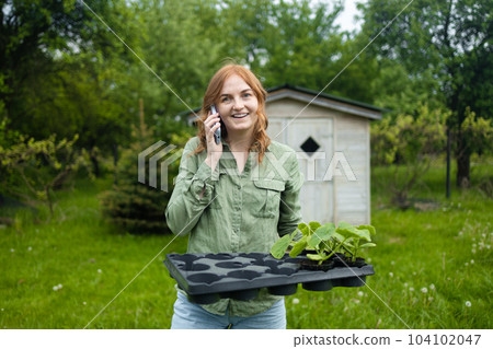 Happy farmer girl using smart phone, holding a little potted cucumber plant. Healthy lifestyle. Gardening. Spring concept, nature and care. 104102047