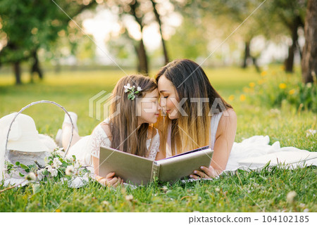 Happy family mom and daughter are watching an album with photos in the summer in the park or in the garden Happy family mom and daughter are watching an album with photos in the summer in the park or in the garden 104102185