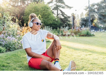 Mixed race young woman with rainbow badge, symbol of LGBTQ social community listening music with wireless headphones while relax in park. Personal Coming out, free lifestyle. Celebrating Pride month 104102385
