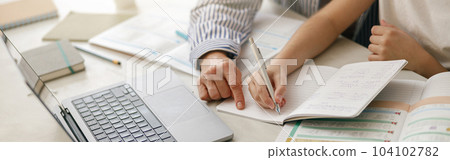 Close up of boy sitting at table and writing in copybook on online lesson while his mom helping him Close up of boy sitting at table and writing in copybook on online lesson while his mom helping him 104102782