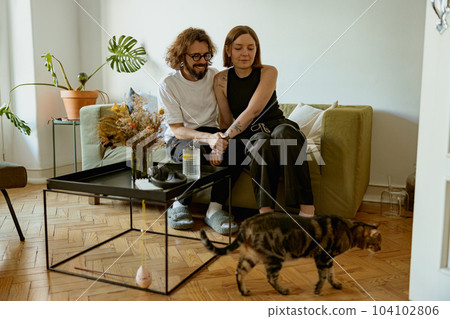Lovely couple sitting on sofa at living room and looking on his domestic cat. Spending time together 104102806