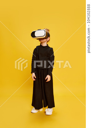 Full-length portrait of little girl, child standing in VR glasses against yellow studio background. Education, fun, game 104102888