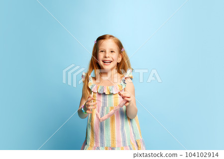 Portrait of beautiful, happy, little girl, child in multicolored striped dress posing with smile and laugh against blue studio background 104102914