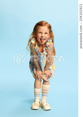 Full-length portrait of little joyful, pretty girl child playing, posing with funny grimacing face against blue studio background 104102921