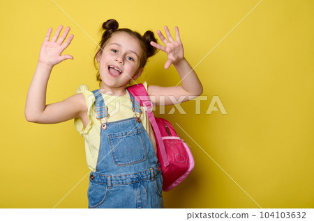 Cheerful preschooler girl showing at camera her hands palms up, smiling, enjoying the start of new school year, posing with pink backpack, dressed in blue denim dress, isolated over yellow background 104103632