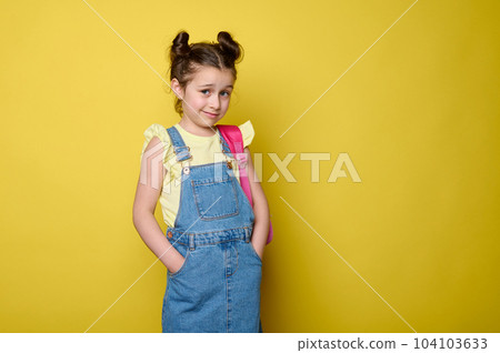 Advertising studio portrait of adorable mischievous little school girl in blue denim overalls, carrying a pink backpack, putting her hands in pockets, looking at camera, isolated on yellow background 104103633