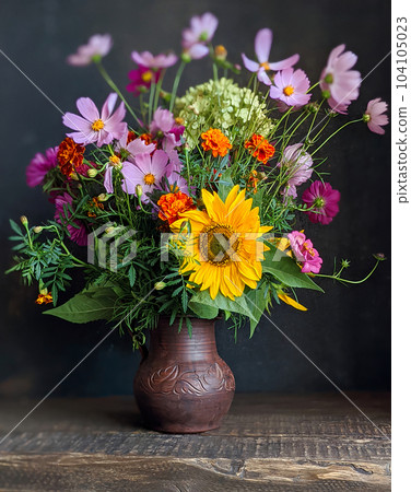 Bouquet of flowers in an old jug on a wooden table with black background. Still life with marigolds, decorative sunflower, kosmeya, hydrangea in the flower arrangement.  104105023