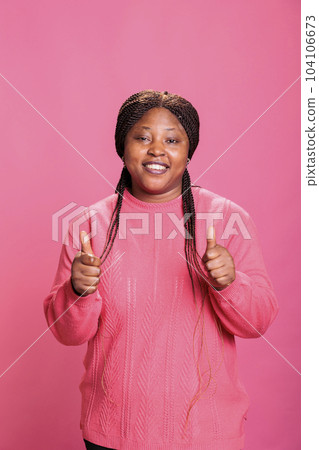 Smiling african american model showing thumbs up sign standing over pink background in studio. Happy cheerful woman with joyful facial expression giving approval gesture, person doing like sign 104106673