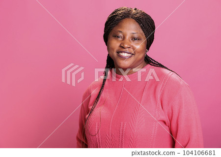 Portrait of african american model smiling at camera while posing happy in studio with pink background, having positive expression. Confident woman with stylish hairstyle and pink sweather 104106681