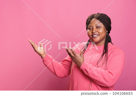 African american promoter pointing to side with palm while advertising product in studio. Cheerful young adut showing promotion standing over pink background, advertise gesture African american promoter pointing to side with palm while advertising product in studio. Cheerful young adut showing promotion standing over pink background, advertise gesture 104106683