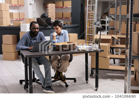 Workers sitting at desk table in storehouse checking shipping logistics on laptop computer before start delivering trendy clothes for clients. Employees working with carton boxes in fashion warehouse Workers sitting at desk table in storehouse checking shipping logistics on laptop computer before start delivering trendy clothes for clients. Employees working with carton boxes in fashion warehouse 104106690