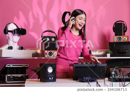 Asian musician with headphones playing electronic song at mixer console, standing at dj table having fun with fans at night in club. Artist performing techno music in studio over pink background 104106713