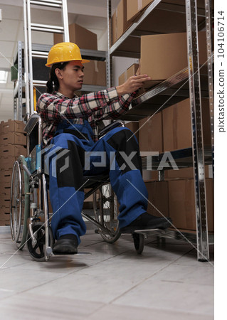 Young asian warehouse worker using wheelchair while doing inventory in storage room. Logistics manager with physical disability checking merchandise parcel on shelf in storehouse 104106714