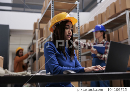 Logistics operator managing stock control on laptop in industrial warehouse. Freight distribution center storehouse employee in yellow hardhat analyzing inventory data on computer Logistics operator managing stock control on laptop in industrial warehouse. Freight distribution center storehouse employee in yellow hardhat analyzing inventory data on computer 104106715