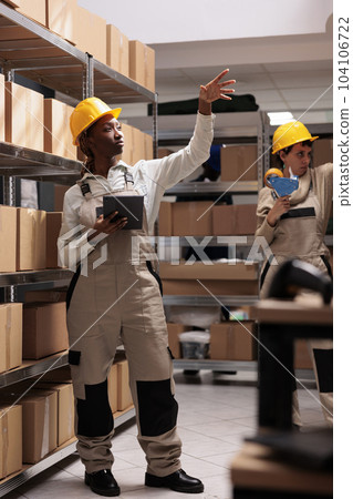 Woman storehouse operator working with digital tablet in warehouse and taking parcel from shelf. African american delivery service storage employee pointing at cardboard boxes 104106722