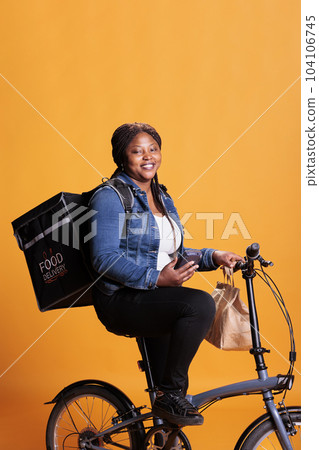 Pizzeria delivery employee looking at customer adreess on smartphone before start delivering fast food order to client. Restaurant worker bringing takeaway meal using bike as transportation Pizzeria delivery employee looking at customer adreess on smartphone before start delivering fast food order to client. Restaurant worker bringing takeaway meal using bike as transportation 104106745