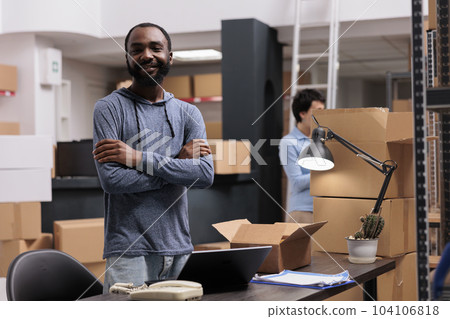 Smiling warehouse employee standing with arm crossed working at clients orders while preparing cardboard boxes in storehouse delivery department. In background woman checking packages shipping details 104106818