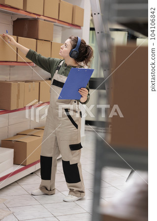 Employee checking carton boxes looking at shipping details, listening music while analyzing clipboard with goods checklist in warehouse. Supervisor working at merchandise delivery in storehouse 104106842