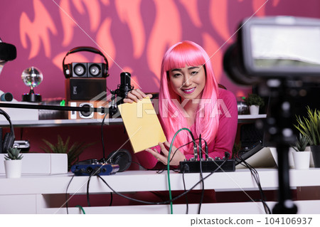 Influencer with pink hair advertising notebook in front of camera recording vlog while presenting school supplies review using professional vlogging equipment. Asian creator filming podcast Influencer with pink hair advertising notebook in front of camera recording vlog while presenting school supplies review using professional vlogging equipment. Asian creator filming podcast 104106937
