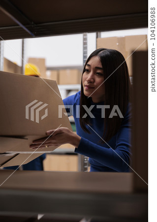Goods supply chain manager doing inventory in warehouse while checking product box on shelf. Asian woman storehouse worker taking freight carton from rack in storage room 104106988