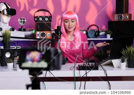 Happy vlogger with pink hair recording podcast while talking with remote subscribers in vlogging studio, posting content on social media. Influencer broadcasting vlog using filming equipment Happy vlogger with pink hair recording podcast while talking with remote subscribers in vlogging studio, posting content on social media. Influencer broadcasting vlog using filming equipment 104106990