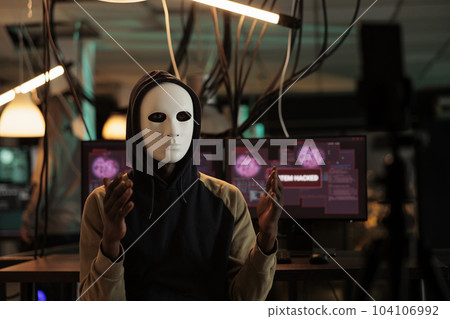 African american hacker broadcasting threat video at night, wearing anonymous mask and asking ransomware. Male spy stealing database information with trojan virus and computer malware. 104106992