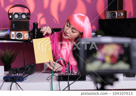 Asian influencer holding notebook advertising product in front of camera while recording school supplies review using broadcast equipment. Content creator with pink hair live streaming Asian influencer holding notebook advertising product in front of camera while recording school supplies review using broadcast equipment. Content creator with pink hair live streaming 104107013