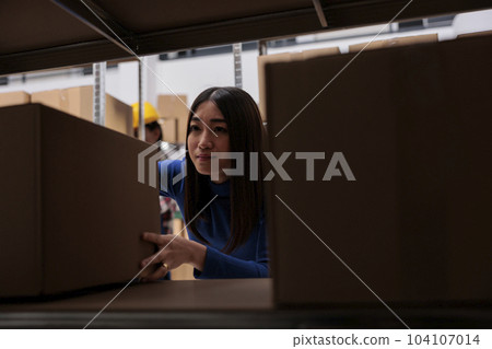 Distribution center worker taking cardboard box from rack to prepare goods for dispatching. Asian woman order picker searching carton package on shelf in industrial warehouse Distribution center worker taking cardboard box from rack to prepare goods for dispatching. Asian woman order picker searching carton package on shelf in industrial warehouse 104107014