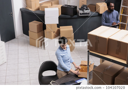 Top view of employee standing at desk in warehouse while preparing client order putting clothes in carton box after checking delivery detalies. Diverse team working in distribution center Top view of employee standing at desk in warehouse while preparing client order putting clothes in carton box after checking delivery detalies. Diverse team working in distribution center 104107020