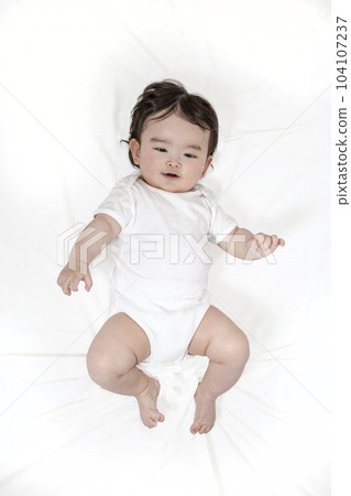Full body figure of an infant looking at the camera while lying on his back on a white sheet bed 104107237