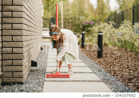 A little girl with a brush cleans a path on the street in the courtyard A little girl with a brush cleans a path on the street in the courtyard 104107692