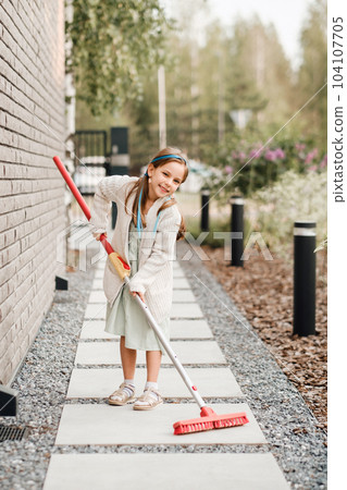 A little girl with a brush cleans a path on the street in the courtyard 104107705